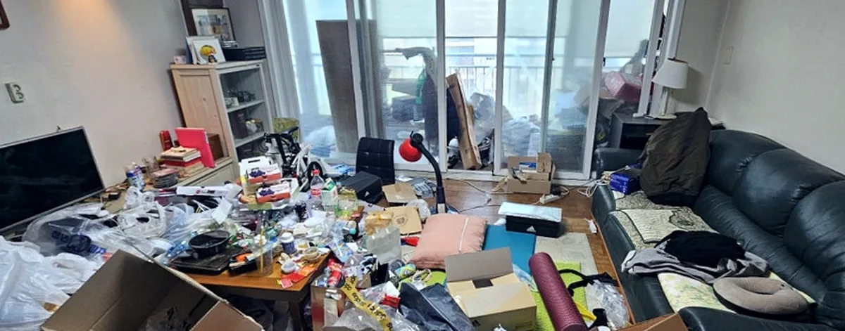 정리 전 거실 — 가죽 소파 위 패딩과 바닥을 덮은 택배상자·비닐봉지