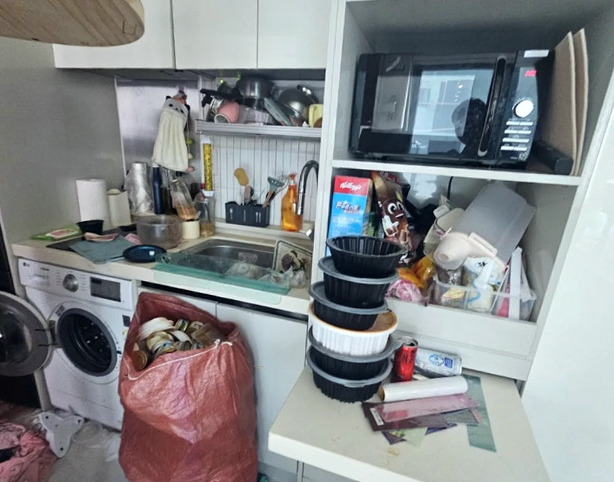 정리 전 주방 — 세탁기 옆 빨간 마대자루에 빈 캔이 가득하고 선반에 배달용 검은 원형 용기가 쌓인 모습