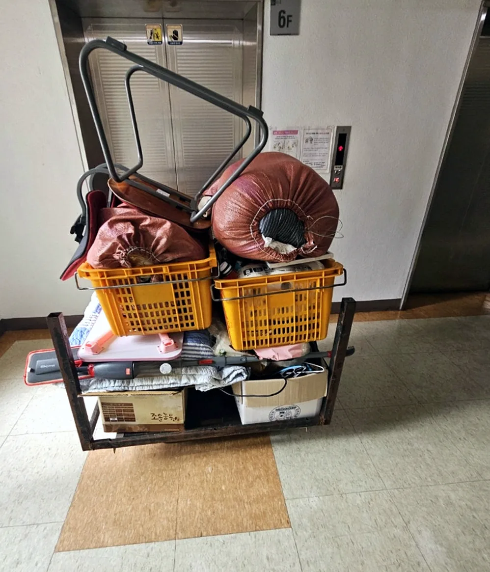 엘리베이터 앞 대차 — 노란 상자 두 개와 묶은 마대, 의자 프레임, 접이식 탁자, 이불을 높이 쌓은 모습